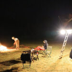Nighttime campfire scene: a person tending the flames while two others sit in folding chairs near a brightly lit vehicle with a ladder nearby.