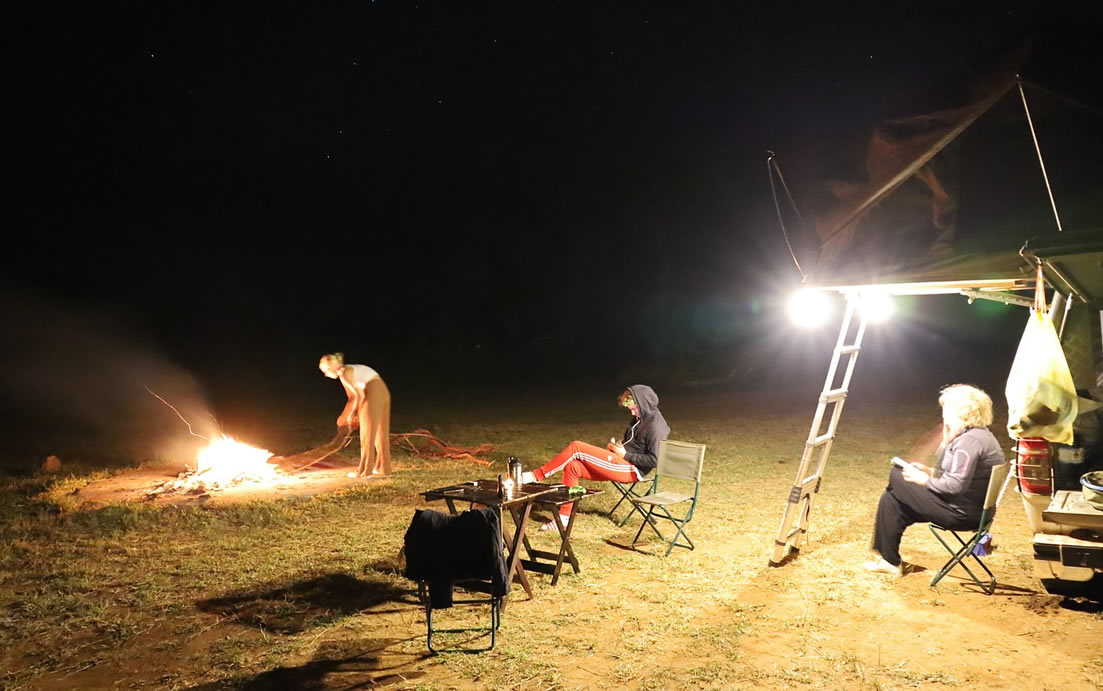 Nighttime campfire scene: a person tending the flames while two others sit in folding chairs near a brightly lit vehicle with a ladder nearby.