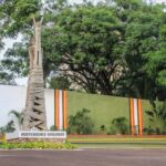 Independence Monument statue wrapped in a spiraled column, on a pedestal with the inscription 'INDEPENDENCE MONUMENT' in a landscaped area with a striped wall behind.