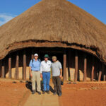 Three men stand in front of a large circular thatched-roof hut with wooden posts on a dirt path in bright sunlight, posing for a photo.