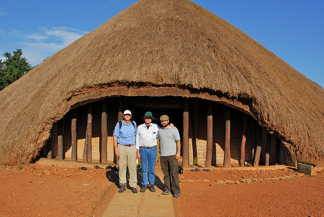 Three men stand in front of a large circular thatched-roof hut with wooden posts on a dirt path in bright sunlight, posing for a photo.