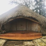 Thatched hut with a curved front porch and bamboo walls set in a garden.