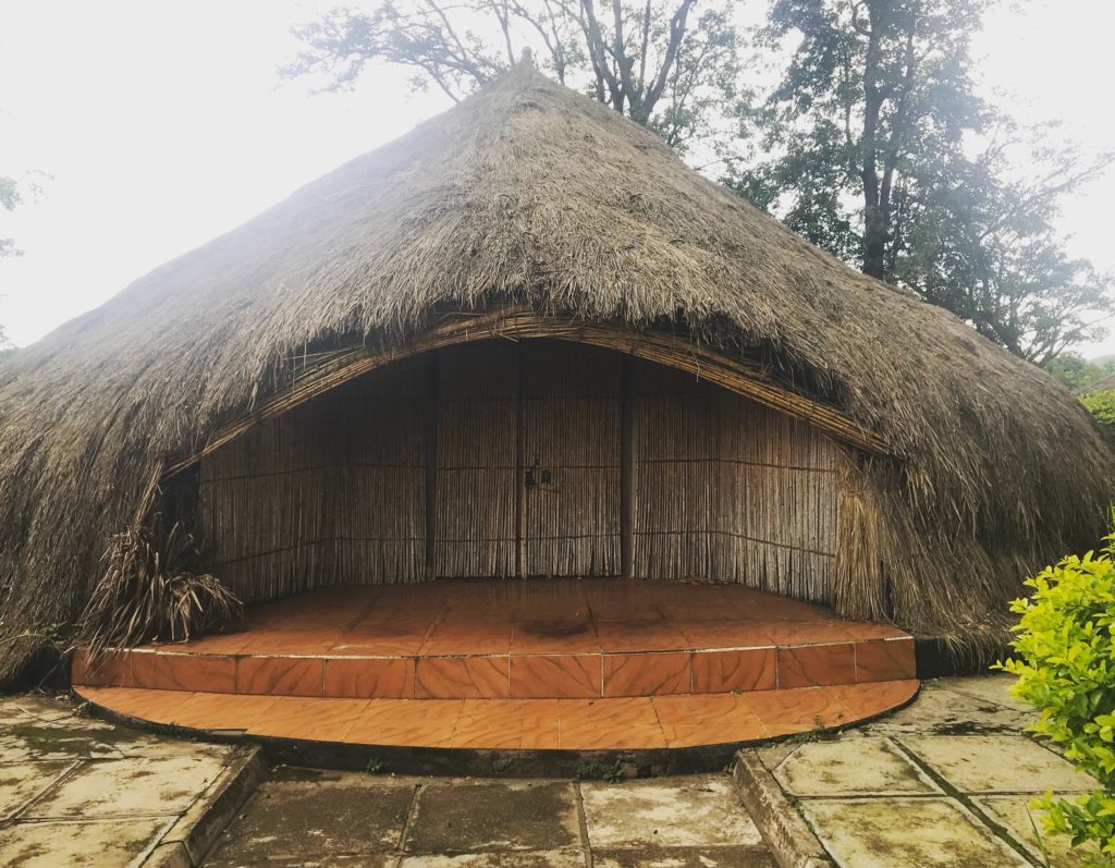 Thatched hut with a curved front porch and bamboo walls set in a garden.