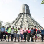 Group of diverse people posing for a photo in front of a large lattice-wrapped tower and surrounding trees on a sunny day.