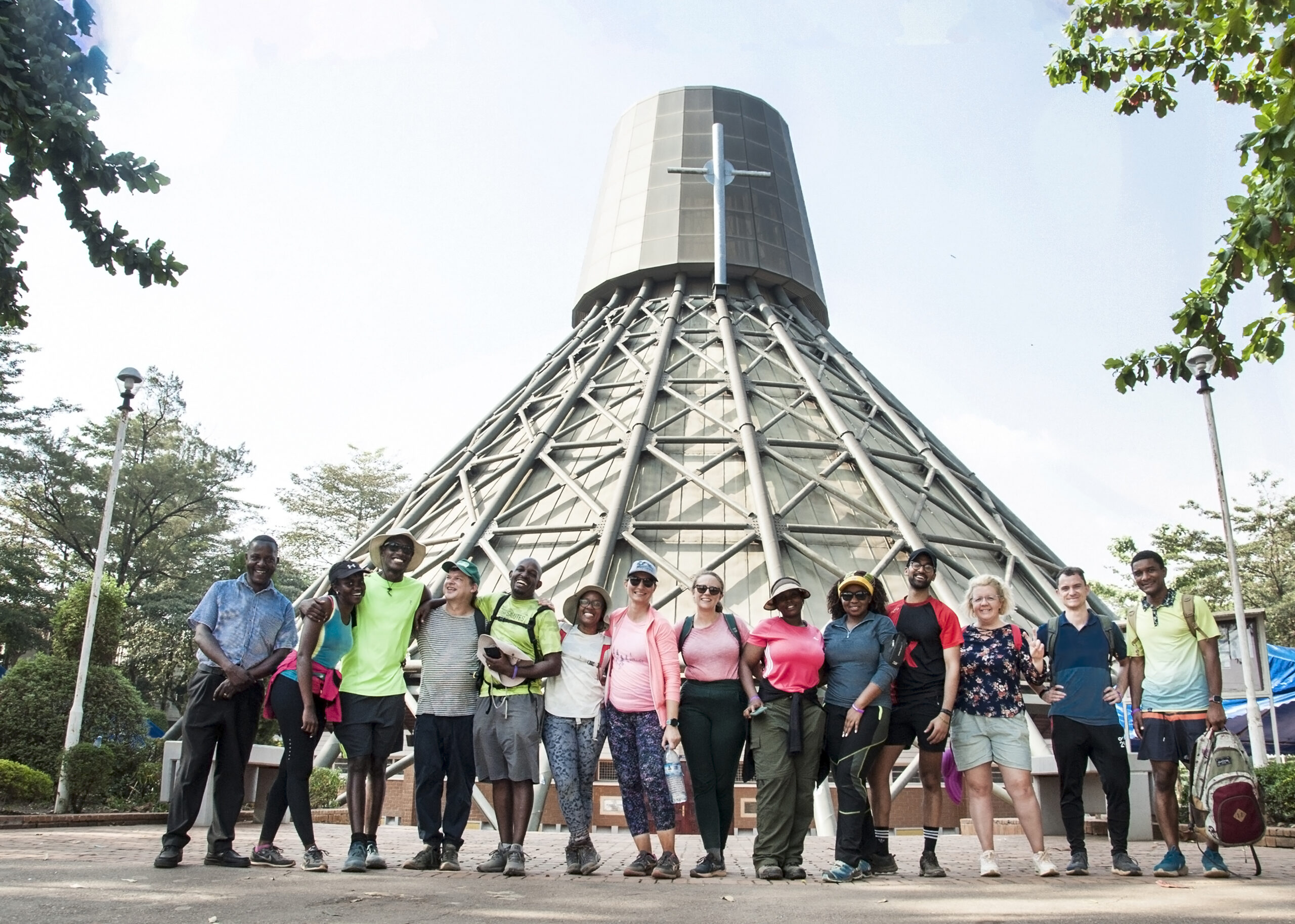 Group of diverse people posing for a photo in front of a large lattice-wrapped tower and surrounding trees on a sunny day.