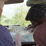 Two men sit in a vehicle examining a folded map spread across the dashboard.