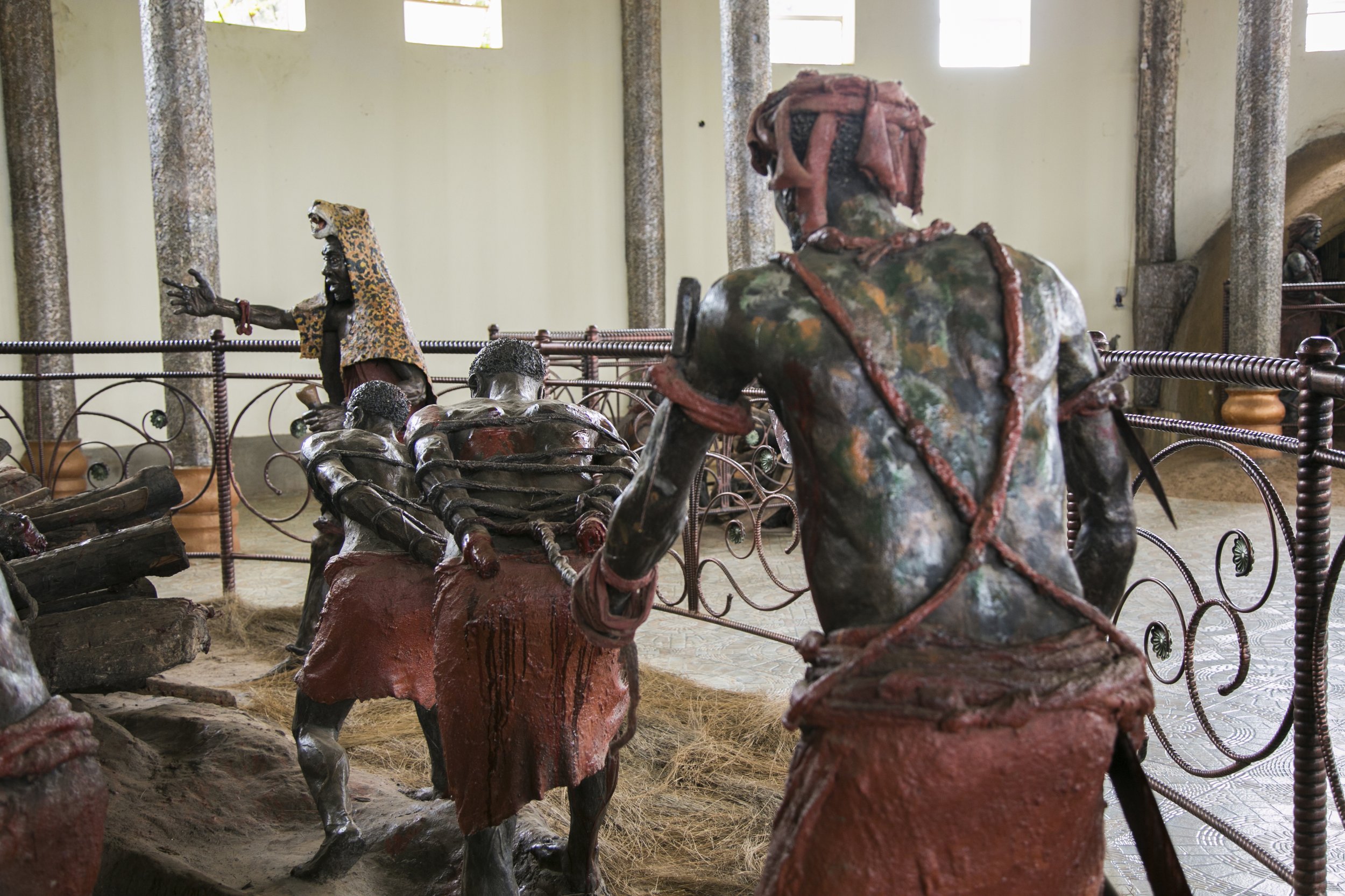 Life-size sculptures of robed figures inside a gallery, some tied with red ropes, surrounded by a metal railing.
