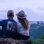 Couple sitting on a grassy hillside, facing distant mountains and valley, arms around each other (back view).
