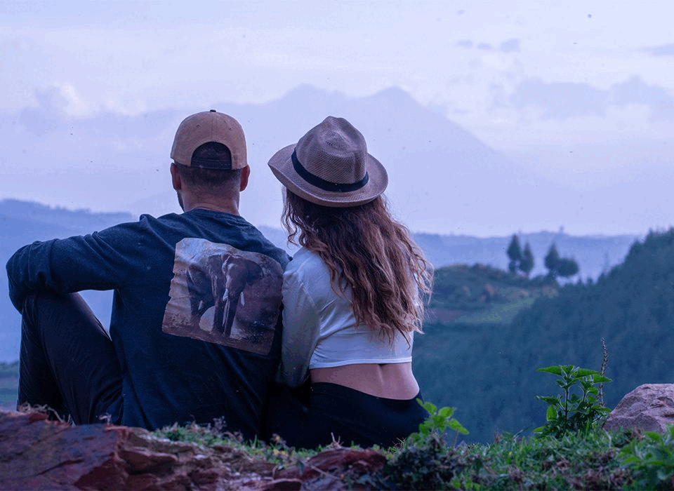 Couple sitting on a grassy hillside, facing distant mountains and valley, arms around each other (back view).