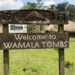 Wooden welcome sign reading 'Welcome to Wamala Tombs' with a thatched hut in a grassy yard behind it.