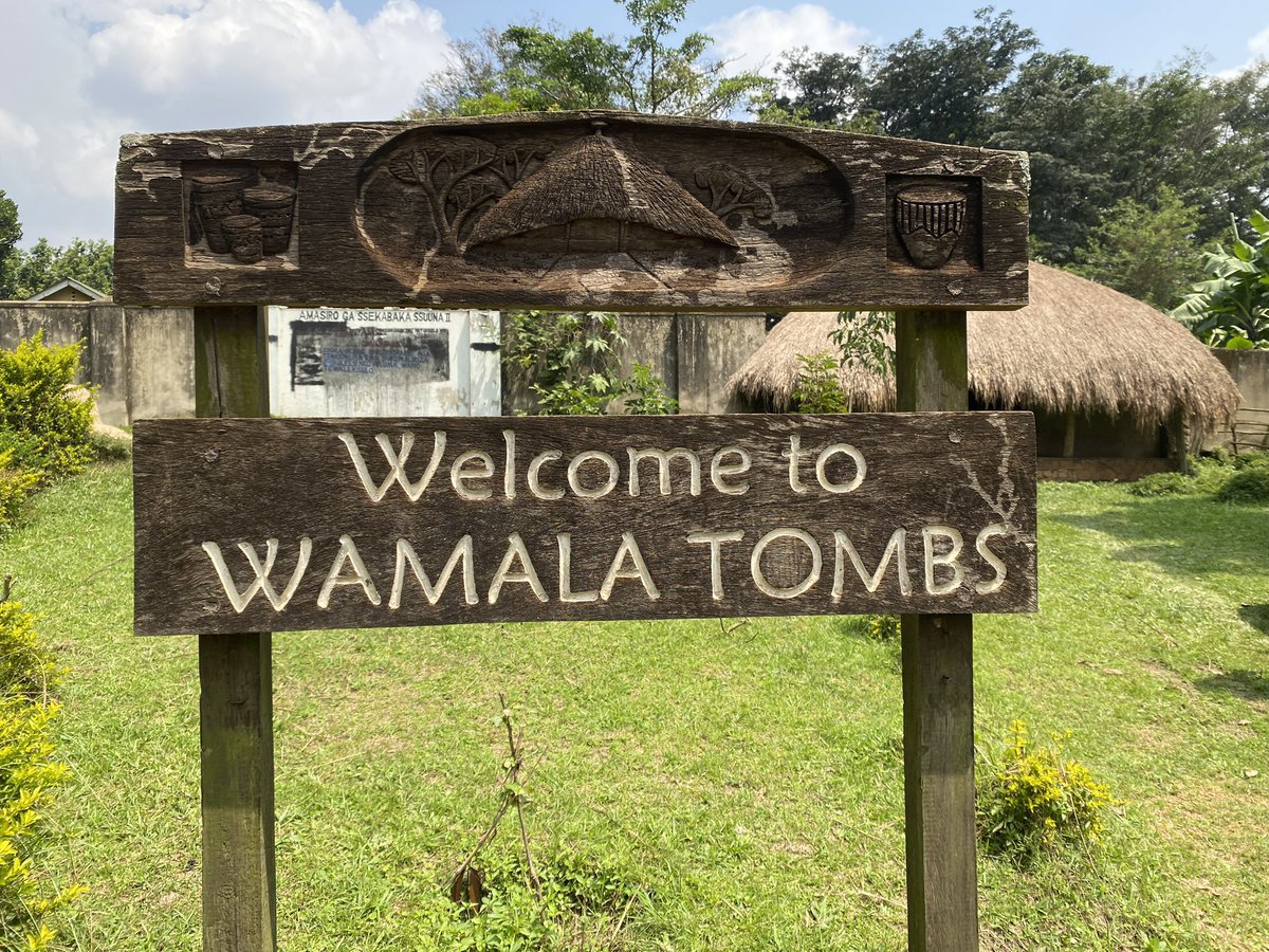 Wooden welcome sign reading 'Welcome to Wamala Tombs' with a thatched hut in a grassy yard behind it.