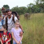 Family of hikers posing in tall grass with a distant elephant in a savanna setting.