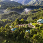 A hillside resort with turquoise-roofed buildings surrounded by dense green forest and misty mountains.